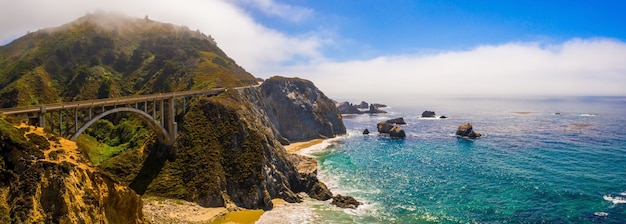 Panoramic aerial shot of the California Bixby bridge on a green hill near beautiful blue water