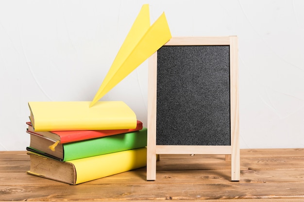 Paper kite on stack of colorful old books and empty blackboard on wooden table 