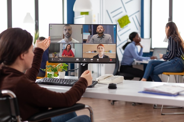 Paralyzed businesswoman in wheelchair discussing marketing strategy with remote team during online videocall meeting conference in business office. Teleconference on computer screen