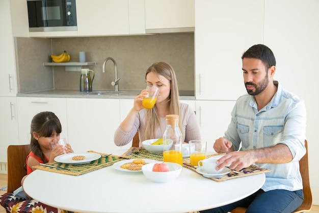 Parents and child having breakfast, drinking orange juice, sitting at dining table with fruits and biscuits.