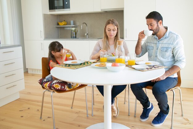 Parents and daughter having breakfast together, drinking coffee and orange juice, sitting at dining table with fruits and biscuits.