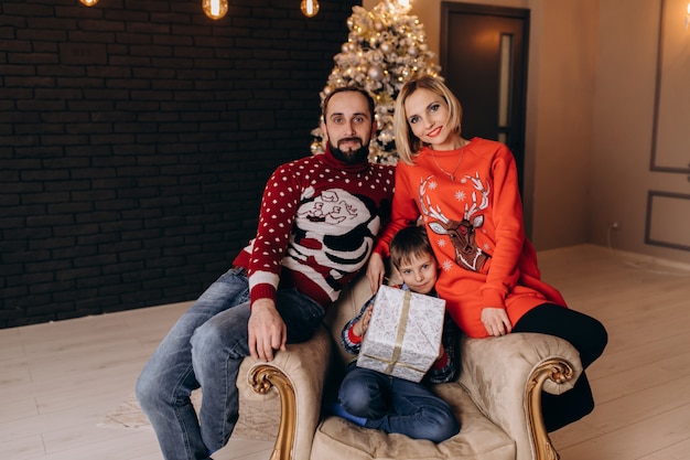 Free photo parents sit around a little boy in soft chair before a christmas tree