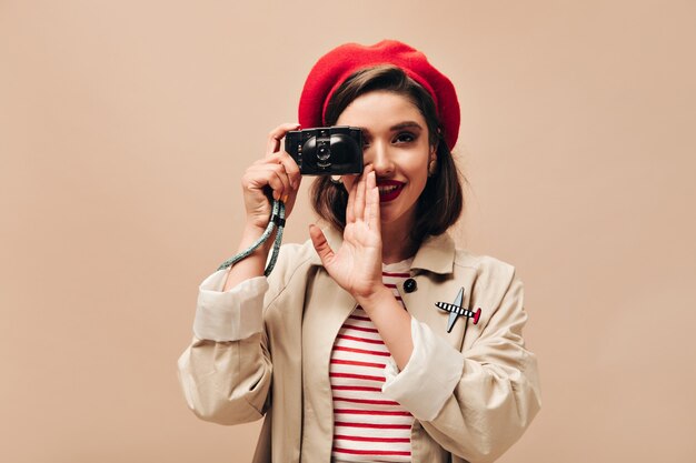 Free photo parisian woman in beret takes photos on beige background. cute young girl with dark hair and bright lips in autumn coat holds black camera.