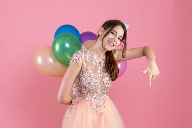 party girl with party cap holding balloons behind her back pointing at down on pink