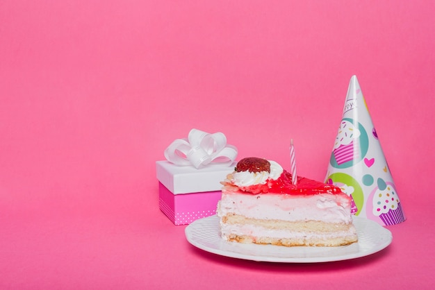 Free photo party hat; gift box and slice of cake with candle on pink background