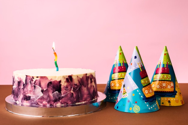 Free photo party hats and cake with lighted candle on desk against pink backdrop