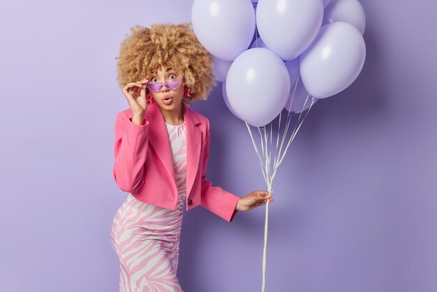 Party time concept Amazed curly haired young woman wears dress has perfect figure stares surprisingly keeps hand on sunglasses holds bunch of inflated balloons ready for birthday celebration