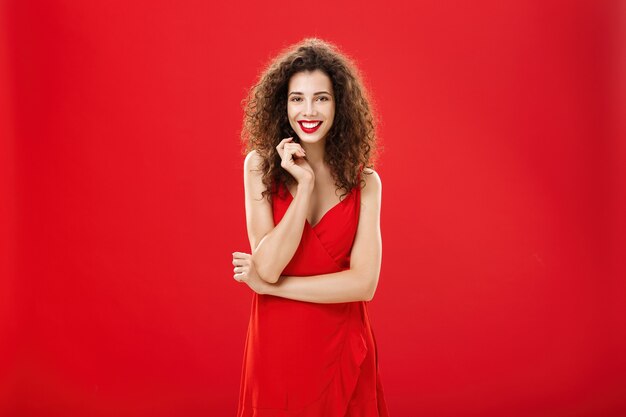 Passionate charming european woman over red background in elegant dress with curly hairstyle smiling cute, feminine playing with hair strand standing timid and silly, talking to person she admires.