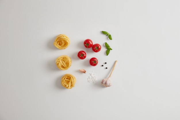 Free photo pasta ingredients on white background. red cherry tomatoes, basil, garlic, peppercorns, uncooked pasta nests for preparing tasty dish. italian cuisine concept. healthy vegetarian diet. flat lay