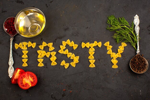 Pasta raw tasty word shaped with oil red sliced tomatoes and greens surrounding on a dark floor