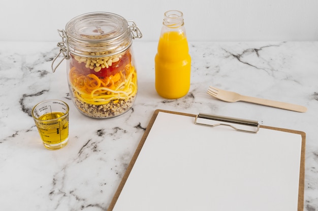 Free photo pasta salad in jar with oil; juice; clipboard and fork on marble surface