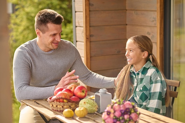 Paternity. Smiling young adult man in casual clothes talking touching shoulder of interestedly listening long haired daughter sitting at table on open veranda on fine afternoon