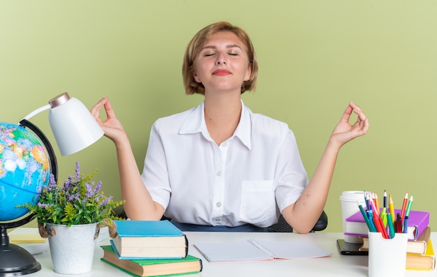 Free Photo peaceful young blonde student girl sitting at desk with school tools meditating with closed eyes isolated on olive green wall