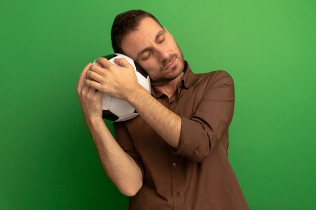 Peaceful young caucasian man holding soccer ball putting head on it with closed eyes isolated on green background with copy space