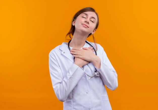 Peaceful young female doctor wearing medical robe and stethoscope putting hands on chest with closed eyes on isolated orange space with copy space