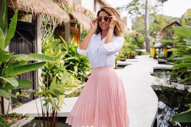 Free photo peaceful young happy woman with short curly hair in pink long skirt and white shirt alone outside her villa