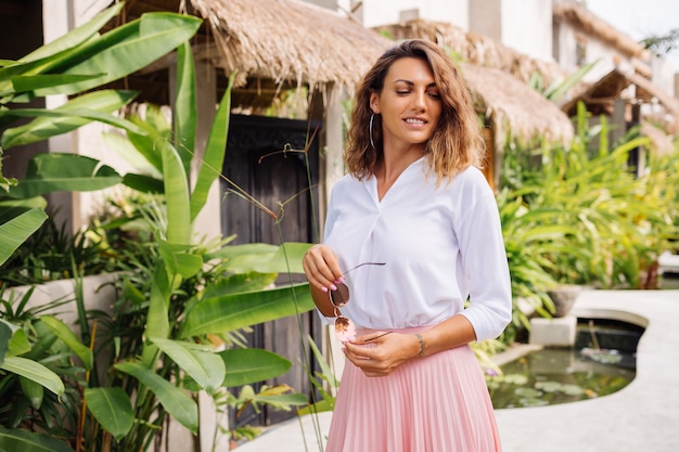 Free photo peaceful young happy woman with short curly hair in pink long skirt and white shirt alone outside her villa