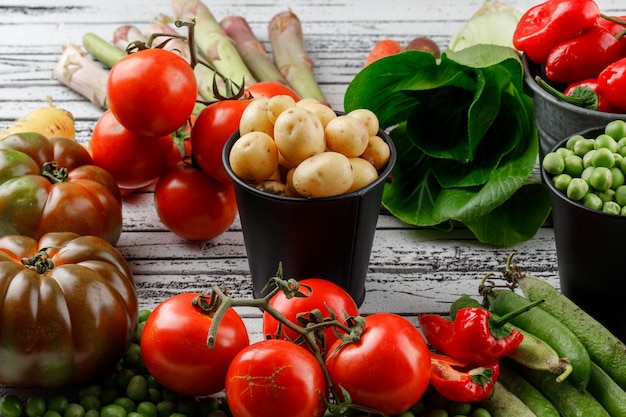 Peas and potatoes with peppers, tomatoes, asparagus, bok choy, green pods, carrots in mini buckets on wooden wall, high angle view.