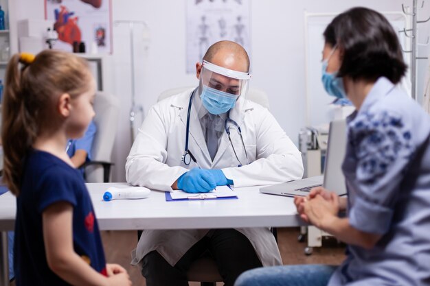 Pediatrician questioning patient about disease and writing answears in clipboard. Medical doctor in protective equipment against coronavirus discussing the symptoms in medical clinic