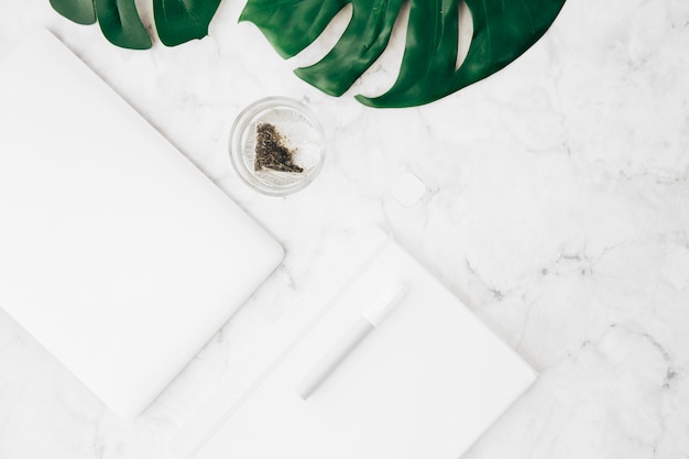 Free Photo pen; diary; digital tablet; monstera leaf and teabag in the glass on marble table