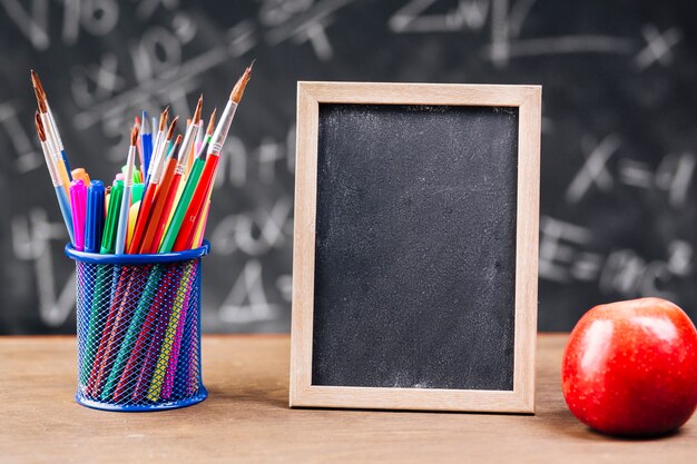 Pen stand and blank chalkboard with apple placed on desk