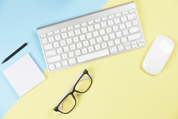 Pencil; adhesive notepad; eyeglasses; wireless keyboard and mouse on blue and yellow backdrop