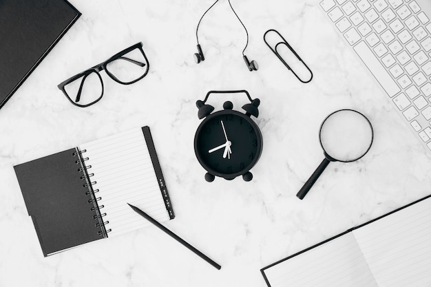 Pencil; diary; eyeglasses; earphone; paperclip; magnifying glasses; alarm clock with keyboard and diary on white desk