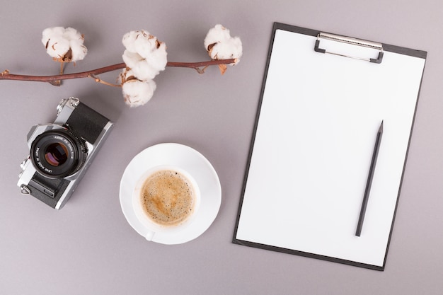 Free photo pencil and paper on clipboard near camera, plant twig and cup