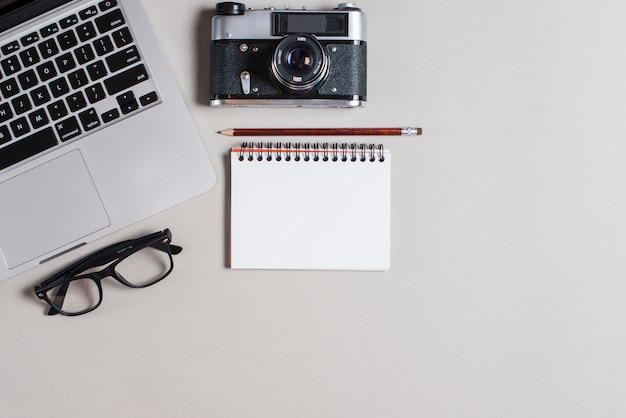 Free photo pencil over spiral notepad with laptop; camera and eyeglasses on gray backdrop