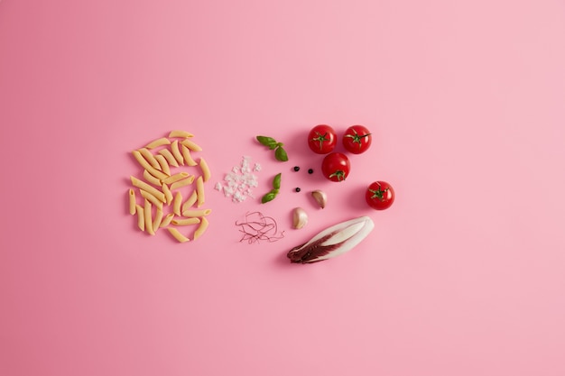 Penne dry rice pasta basil, chicory salad, tomatoes, garlic red chili pepper threads for preparing delicious gourmet Italian cuisine. Uncooked macaroni and ingredients on rosy background. Healthy food