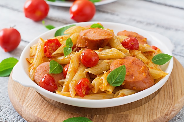 Free photo penne pasta with tomato sauce with sausage, tomatoes, green basil decorated in a frying pan on a wooden table