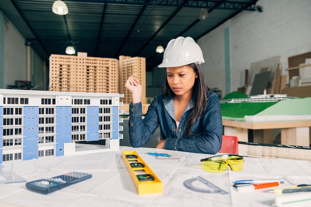 Pensive African-American woman in safety helmet near model of building