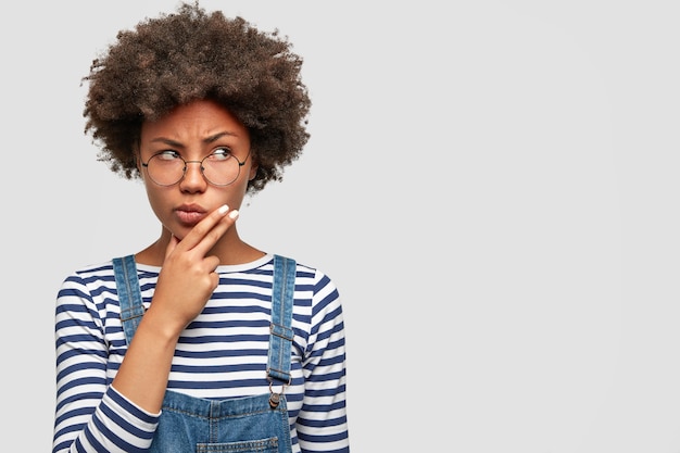 Pensive African dark skinned female holds chin and looks thoughtfully aside