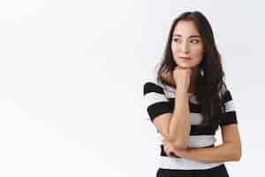 Free photo pensive, attractive and thoughtful young asian brunette woman in striped t-shirt, hold hand under chin, looking away dreamy, smiling contemplating someone passing by, standing white background