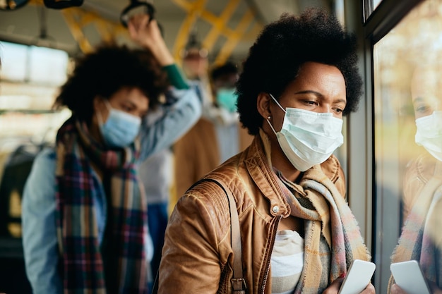 Pensive black woman with face mask commuting by bus and looking through the window