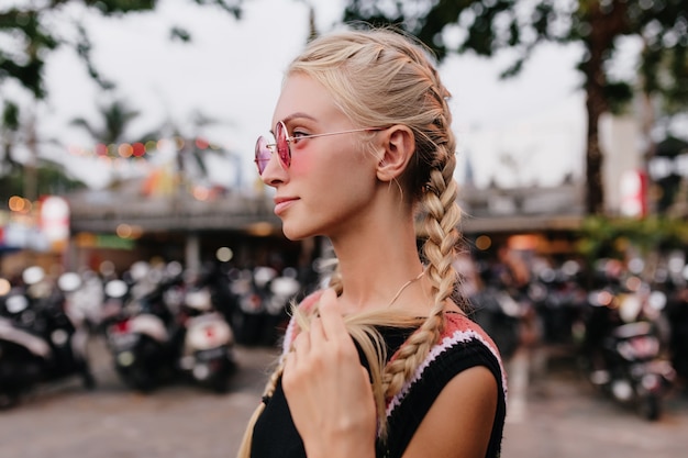 Free photo pensive blonde woman in black attire posing on blur street background. tanned lady with braids wears pink sunglasses.