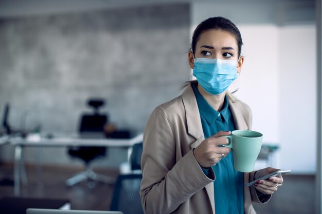 Pensive businesswoman with face mask using cell phone on coffee break in the office