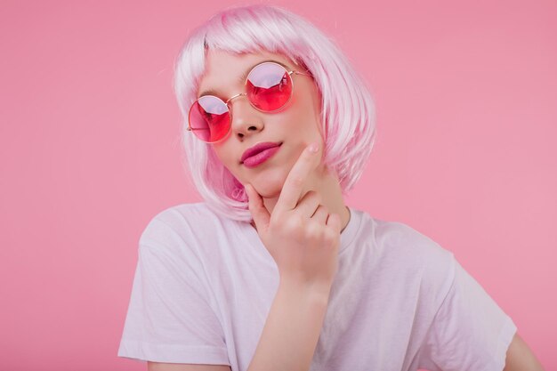 Pensive charming girl with pink hair looking up while posing on pastel background Carefree european woman in sunglasses and periwig chilling during studio photoshoot