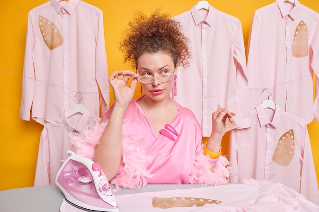 Free photo pensive curly haired woman keeps hand on rim of spectacles dressed in silk gown poses near ironing board with electric iron poses against shirts on hangers thinks how to finish domestic work in time