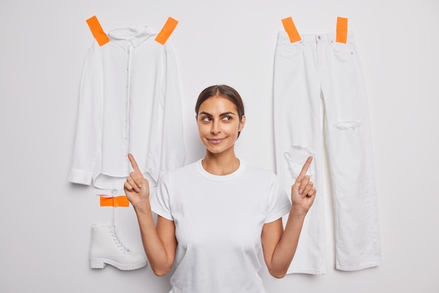 Pensive dark haired pretty woman points index fingers above advertises clothes isolated over white background with plastered t shirt trousers and boot Monochrome shot You should wear this