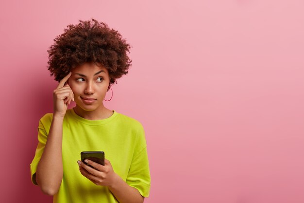 Pensive dark skinned woman keeps finger on temple, looks aside thoughtfully, uses new gadget for first time, dressed casually, tries to remember name of product before browsing internet, has idea