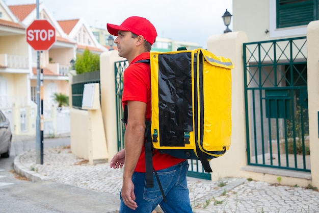 Free photo pensive deliveryman in red cap watching required house. middle-aged courier with yellow thermal backpack delivering express order and walking on street. delivery service and online shopping concept