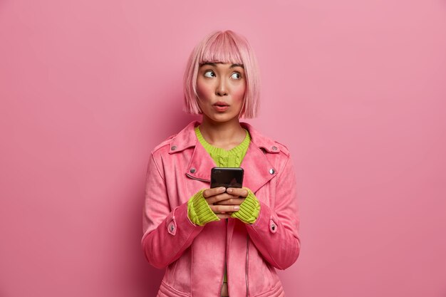 Pensive doubtful woman with bob hairstyle, browses newsfeed via social networks, looks away thoughtfully with concentrated expression