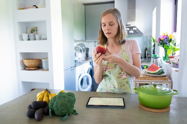 Free photo pensive focused woman holding fruit while cooking in kitchen, using tablet near saucepan and fresh vegetables on counter. front view. cooking at home and healthy eating concept