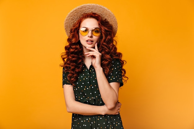 Pensive ginger girl posing in sunglasses and straw hat. Front view of romantic european lady isolated on yellow background.