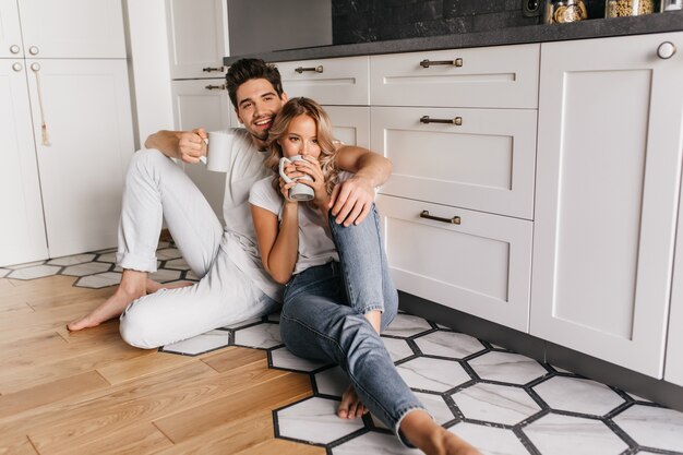 Pensive girl sitting on the floor with cup of coffee. Happy couple enjoying tea together.