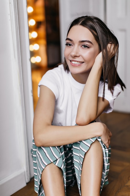 Pensive girl with big dark eyes sitting on the floor. Smiling beautiful lady in pajamas spending weekend in her apartment.