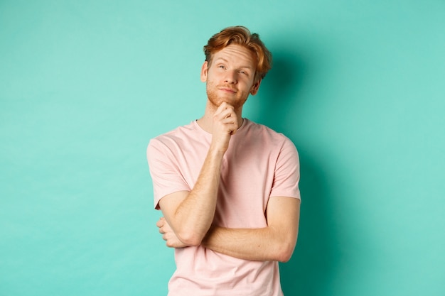 Pensive handsome man with red hair and beard looking at upper left corner, making choice and looking thoughtful, standing in t-shirt over mint background.