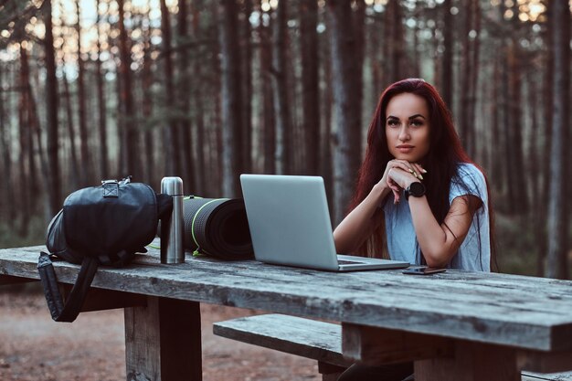 Free Photo pensive hipster girl in white shirt looking at camera while sitting on a wooden bench with an open laptop in a beautiful autumn forest.