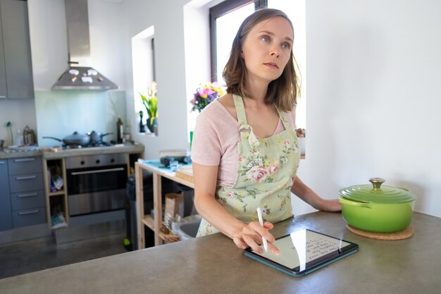 Free photo pensive housewife planning weekly menu and writing grocery list in her kitchen, using tablet near big saucepan on counter, looking up. cooking at home and online cookbook concept